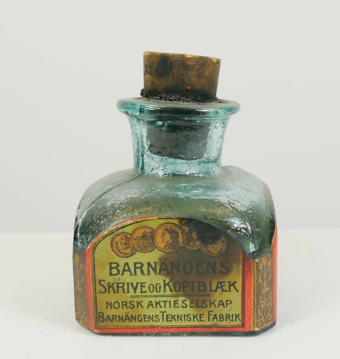 Kvadratisk glassflaske til blekk. Flasken smalner i en tut øverst og er tettet med en kork. Rundt sidene er det papiretiketter. 