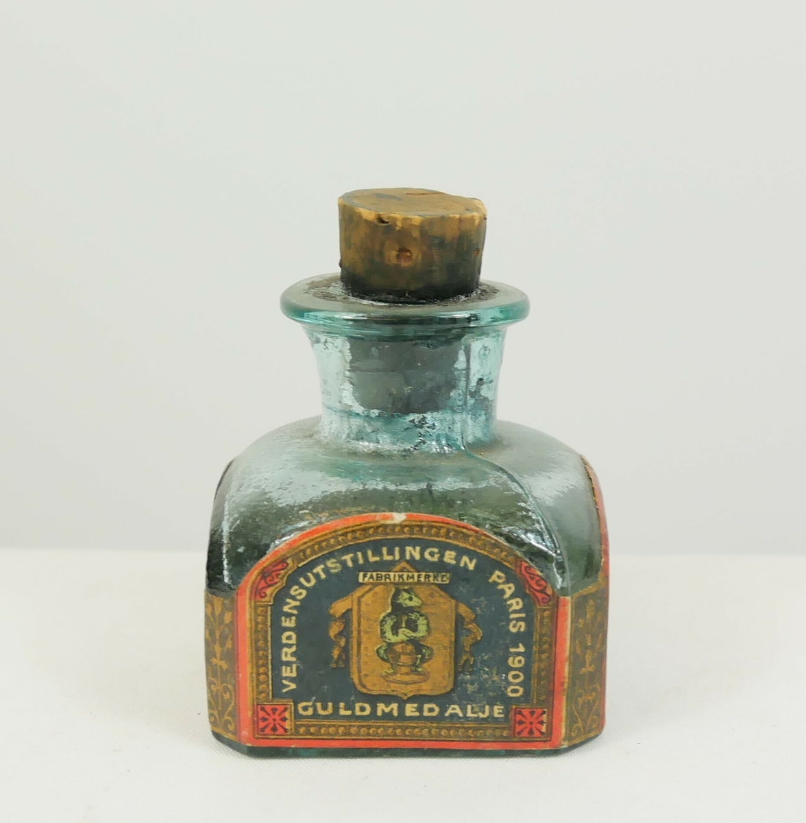 Kvadratisk glassflaske til blekk. Flasken smalner i en tut øverst og er tettet med en kork. Rundt sidene er det papiretiketter. 