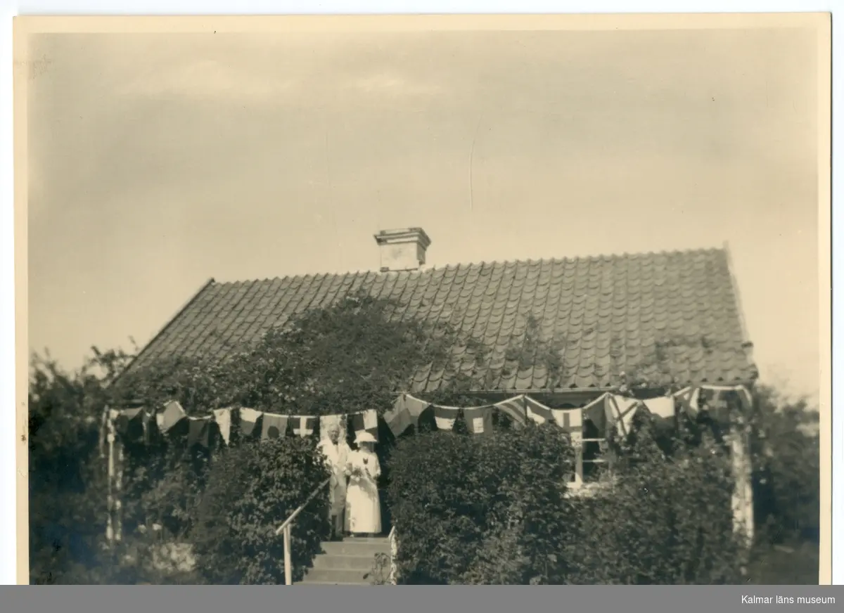 Svart-vitt fotografi. Waldemar Swahn och en kvinna står vitklädda på trappan till Waldemar Swahns sommarboställe på Stångudden, Ekenäs. Längs hustaket hänger flaggor på rad.