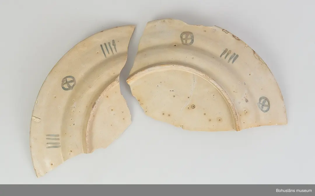 Fat med dekor, del av. Ljusgult-beige skärv. Smal rel. hög fotring. Brättesbredd: 4,5 cm. Färg: Mangan grund. Motiv: på brättet två större fiskar, på spegel; överkroppar/ansikten av två män, en med metspö och fisk, en med vingar på rygg (ängel?). Undersida brätte är dekorerat med cirkelkryss varvat med fyrstreck. Samhör och har passning med fat fnr: 1333A.
Datering: ca 1735-1750 ? Delftgods troligen av engelskt ursprung.