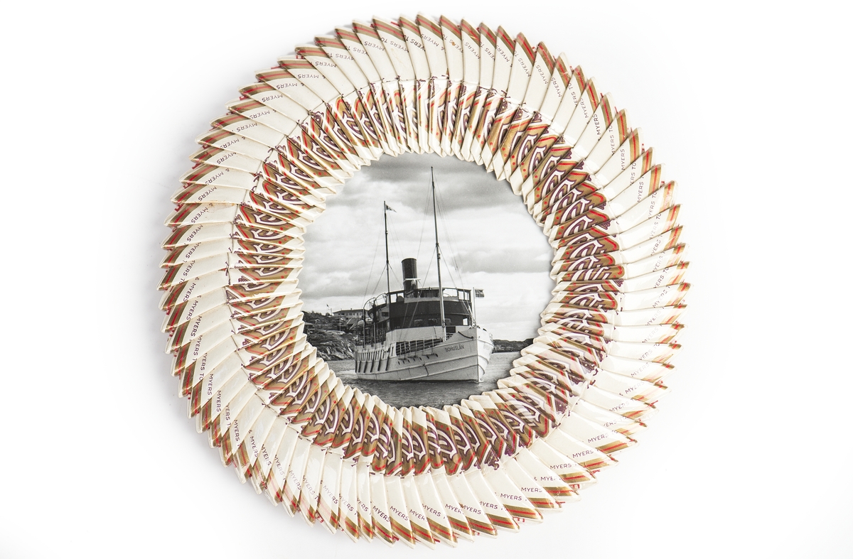 Inramat s/v fotografi av Ångaren "Bohuslän”.
Den runda ramen utgörs av vikta cigarettpaket.