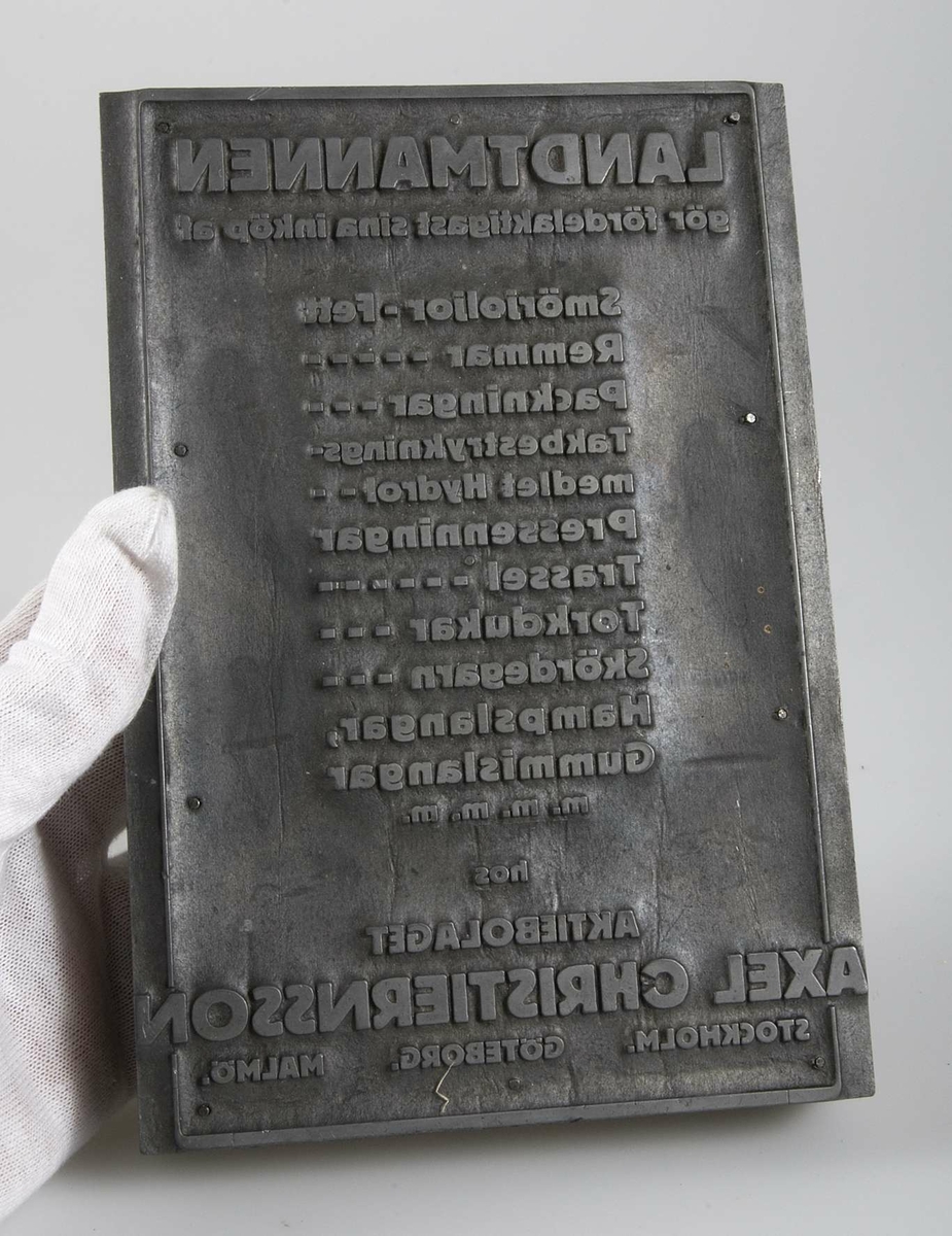 Kliché av bly på rektangulär träplatta. Text LANDTMÄNNEN gör fördelaktigast sina inköp av Smörjolja, Remmar, Packningar, Takbestrykningsmedel ... hos Aktiebolaget AXEL CHRISTIERNSSON, Stockholm, Malmö, Göteborg.