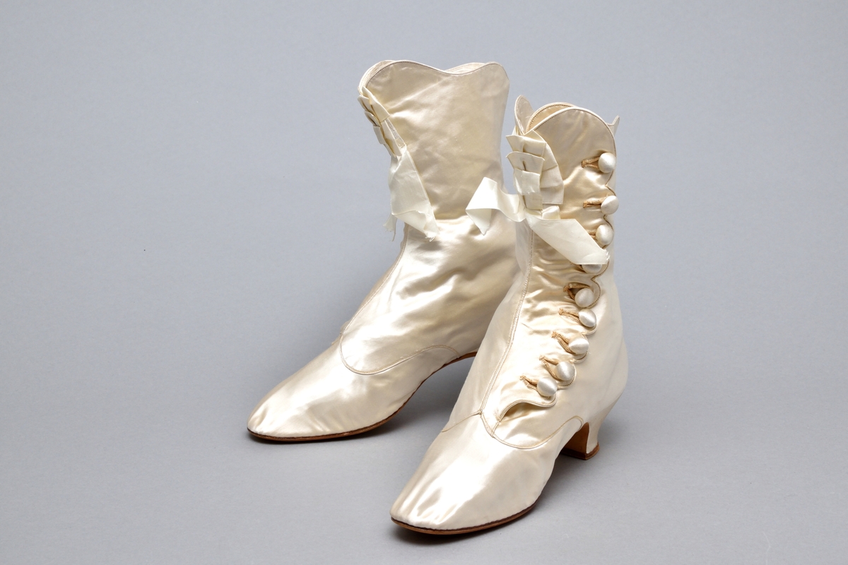Ett par brudkängor av vitt siden tillhörande bruddräkt. Klädd insvängd klack, ca. 3 cm hög. Knäppning med 9 sidenklädda knappar och knapphål längs vågskuren kant på skons utsida. Skaftet med böljande kant. Mitt fram rosett av sidenband. Vitt foder av siden. Lädersula stämplad: PINET, Paris.