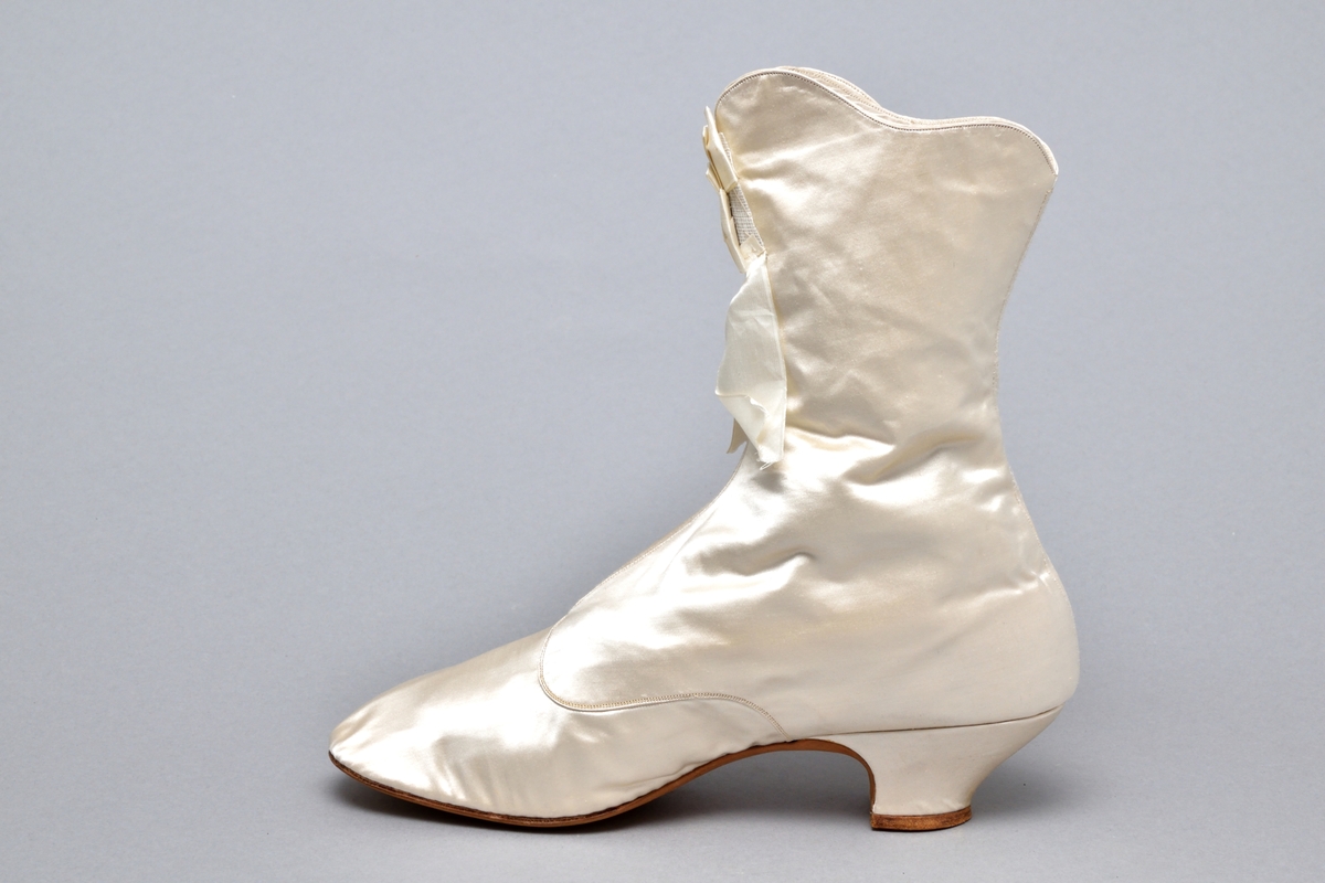 Ett par brudkängor av vitt siden tillhörande bruddräkt. Klädd insvängd klack, ca. 3 cm hög. Knäppning med 9 sidenklädda knappar och knapphål längs vågskuren kant på skons utsida. Skaftet med böljande kant. Mitt fram rosett av sidenband. Vitt foder av siden. Lädersula stämplad: PINET, Paris.