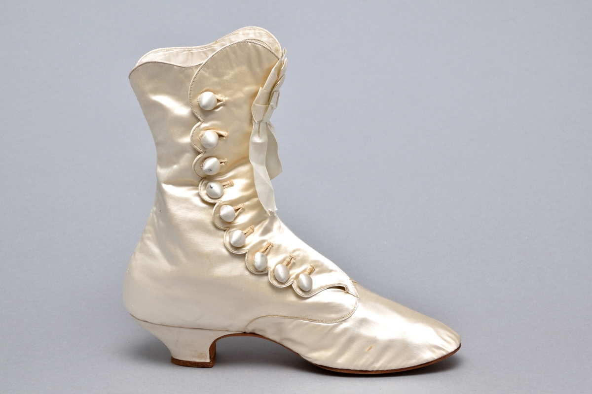 Ett par brudkängor av vitt siden tillhörande bruddräkt. Klädd insvängd klack, ca. 3 cm hög. Knäppning med 9 sidenklädda knappar och knapphål längs vågskuren kant på skons utsida. Skaftet med böljande kant. Mitt fram rosett av sidenband. Vitt foder av siden. Lädersula stämplad: PINET, Paris.