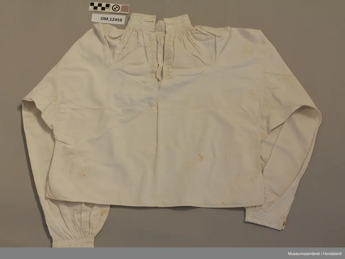 Skjorte med splitt framme og broderi i halsen og langs splitten og framme på ermene. Kvitsaum med mange primhol. Halsen og ermlinningane er forte med lin. Sjølve skjorta er i bomullslerret. Splitt i sidene og kiler under ermene og opp på skuldrane. Knapphol framme på halslinningen på begge sider. Krok og malje i ermlinningane og halslinningen. Broderiet er svært slitt. Litt flekkete.