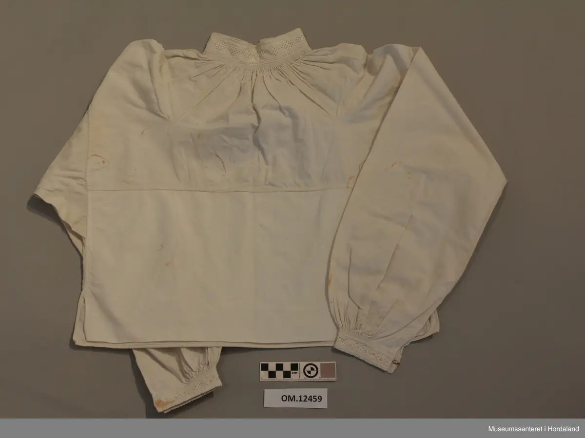 Skjorte med splitt framme og broderi i halsen og langs splitten og framme på ermene. Kvitsaum med mange primhol. Halsen og ermlinningane er forte med lin. Sjølve skjorta er i bomullslerret. Splitt i sidene og kiler under ermene og opp på skuldrane. Knapphol framme på halslinningen på begge sider. Krok og malje i ermlinningane og halslinningen. Broderiet er svært slitt. Litt flekkete.