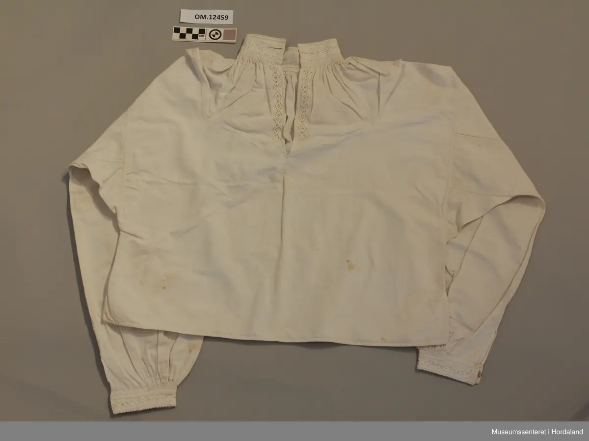 Skjorte med splitt framme og broderi i halsen og langs splitten og framme på ermene. Kvitsaum med mange primhol. Halsen og ermlinningane er forte med lin. Sjølve skjorta er i bomullslerret. Splitt i sidene og kiler under ermene og opp på skuldrane. Knapphol framme på halslinningen på begge sider. Krok og malje i ermlinningane og halslinningen. Broderiet er svært slitt. Litt flekkete.