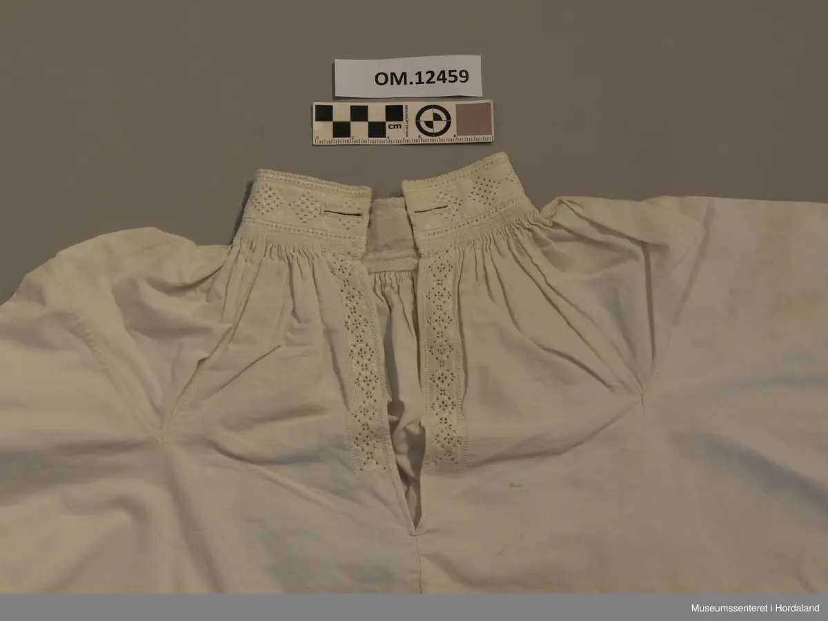 Skjorte med splitt framme og broderi i halsen og langs splitten og framme på ermene. Kvitsaum med mange primhol. Halsen og ermlinningane er forte med lin. Sjølve skjorta er i bomullslerret. Splitt i sidene og kiler under ermene og opp på skuldrane. Knapphol framme på halslinningen på begge sider. Krok og malje i ermlinningane og halslinningen. Broderiet er svært slitt. Litt flekkete.