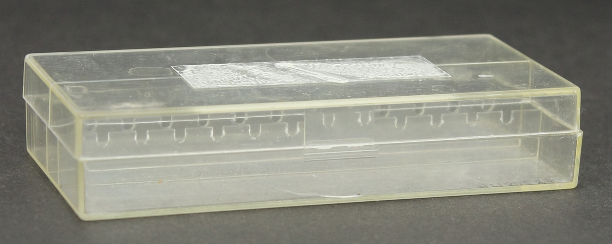 Rektangulär ask med lock av transparent plast. Invändigt i askens mitt finns två parallella sarger med 12 U-formade urgröpningar i vardera, har innehållit ett dussin hålborstar. Asken har gångjärn i plast på bakkanten ett av två har gått av därav sitter inte locket fast. Asken är repig och har en spricka i framkant samt en brunaktig fläck i bakkant. Locket har en sarg i mitten invändigt. Utanpå locket finns en rektangulär fort, troligtvis har det tidigare varit en etikett där nu är det färgad/målad flammigt vit.