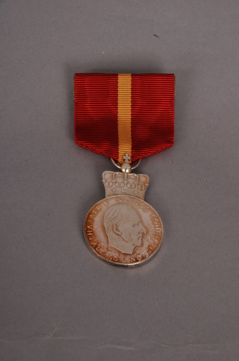 Kongens fortjenestmedalje i sylv, med etui. Etuiet er i raud plast, med fløyel inni. Medalja er sylvfarga, med raudt oppheng med gul stripe.