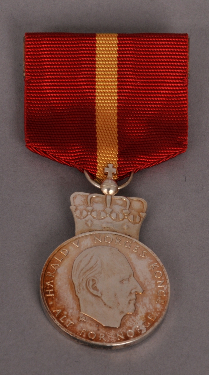Kongens fortjenestmedalje i sylv, med etui. Etuiet er i raud plast, med fløyel inni. Medalja er sylvfarga, med raudt oppheng med gul stripe.