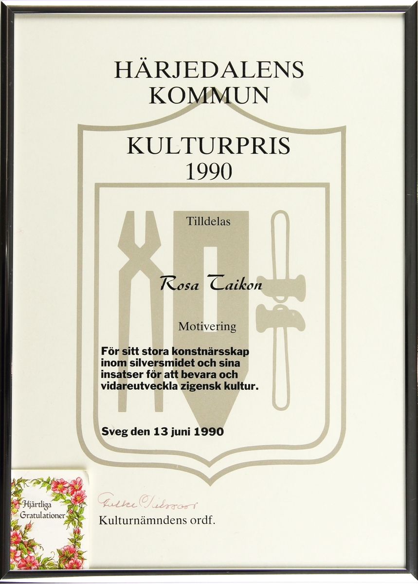 Diplom för Kulturpris från Härjedalens kommun till Rosa Taikon. Diplomet är av vitt papper med kommunvapnet tryckt i grått i bakgrunden. Texten är i svart och diplomet är inramat i en metallisk ram. I det nedre vänstra hörnet sitter en kort med blomdekoration.