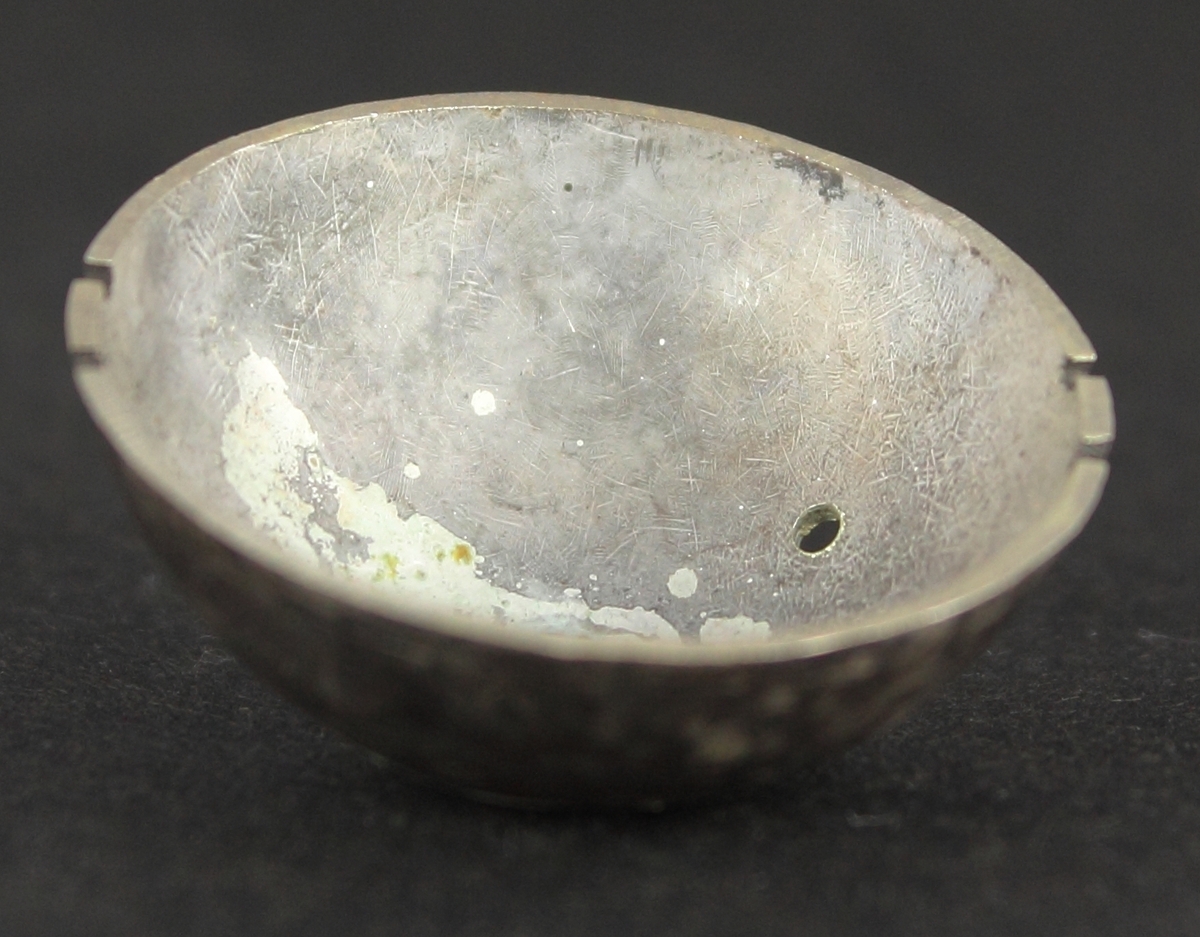 En silverplåt formad rund och skålformad. Silverplåten har en knopp upptill och hål på sidan. Fyra fördjupningar är gjorda längs kanten. Plåten är kraftigt oxiderad.