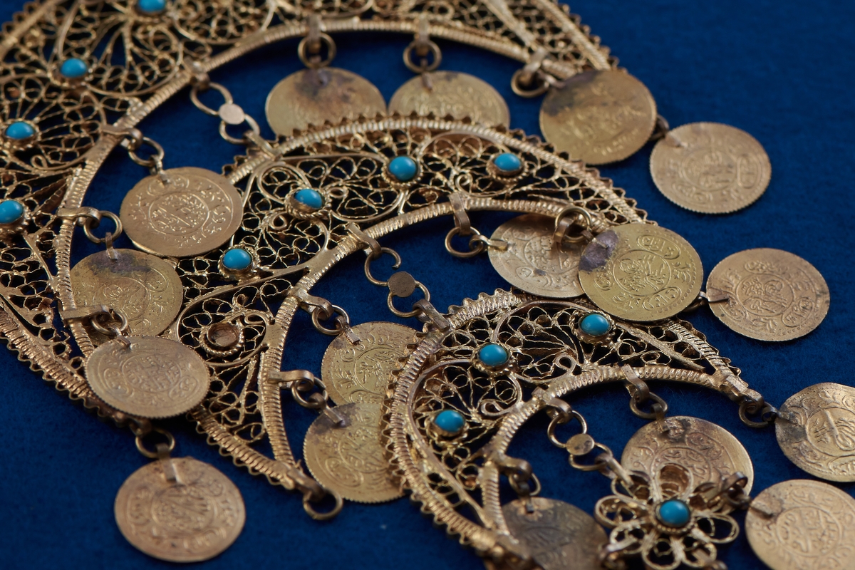 Del av halssmycke, bestående av metallbitar i form av tre halvmånar. Smycket är skapat i gul metall med infattade turkosfärgade stenar. Från varje halvmåne hänger cirklar i bindöglor. Smycket är troligen inköpt av Rosa Taikon vid en resa utomlands, och hängde senare på anslagstavlan i hennes ateljé.