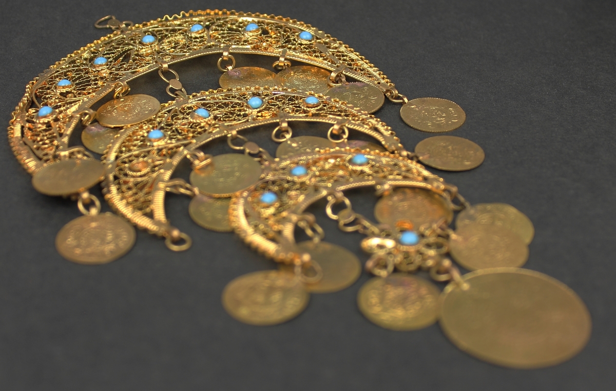 Del av halssmycke, bestående av metallbitar i form av tre halvmånar. Smycket är skapat i gul metall med infattade turkosfärgade stenar. Från varje halvmåne hänger cirklar i bindöglor. Smycket är troligen inköpt av Rosa Taikon vid en resa utomlands, och hängde senare på anslagstavlan i hennes ateljé.