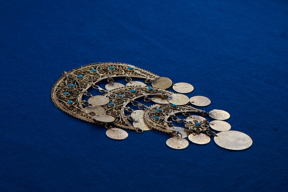 Del av halssmycke, bestående av metallbitar i form av tre halvmånar. Smycket är skapat i gul metall med infattade turkosfärgade stenar. Från varje halvmåne hänger cirklar i bindöglor. Smycket är troligen inköpt av Rosa Taikon vid en resa utomlands, och hängde senare på anslagstavlan i hennes ateljé.