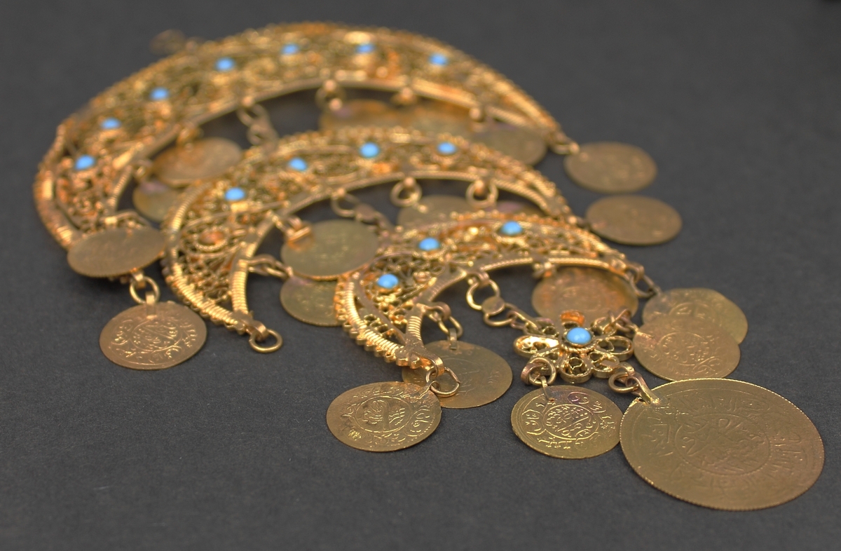 Del av halssmycke, bestående av metallbitar i form av tre halvmånar. Smycket är skapat i gul metall med infattade turkosfärgade stenar. Från varje halvmåne hänger cirklar i bindöglor. Smycket är troligen inköpt av Rosa Taikon vid en resa utomlands, och hängde senare på anslagstavlan i hennes ateljé.