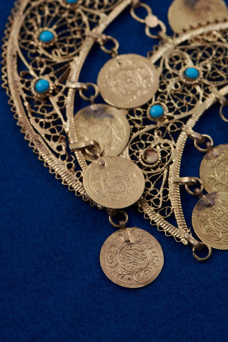 Del av halssmycke, bestående av metallbitar i form av tre halvmånar. Smycket är skapat i gul metall med infattade turkosfärgade stenar. Från varje halvmåne hänger cirklar i bindöglor. Smycket är troligen inköpt av Rosa Taikon vid en resa utomlands, och hängde senare på anslagstavlan i hennes ateljé.