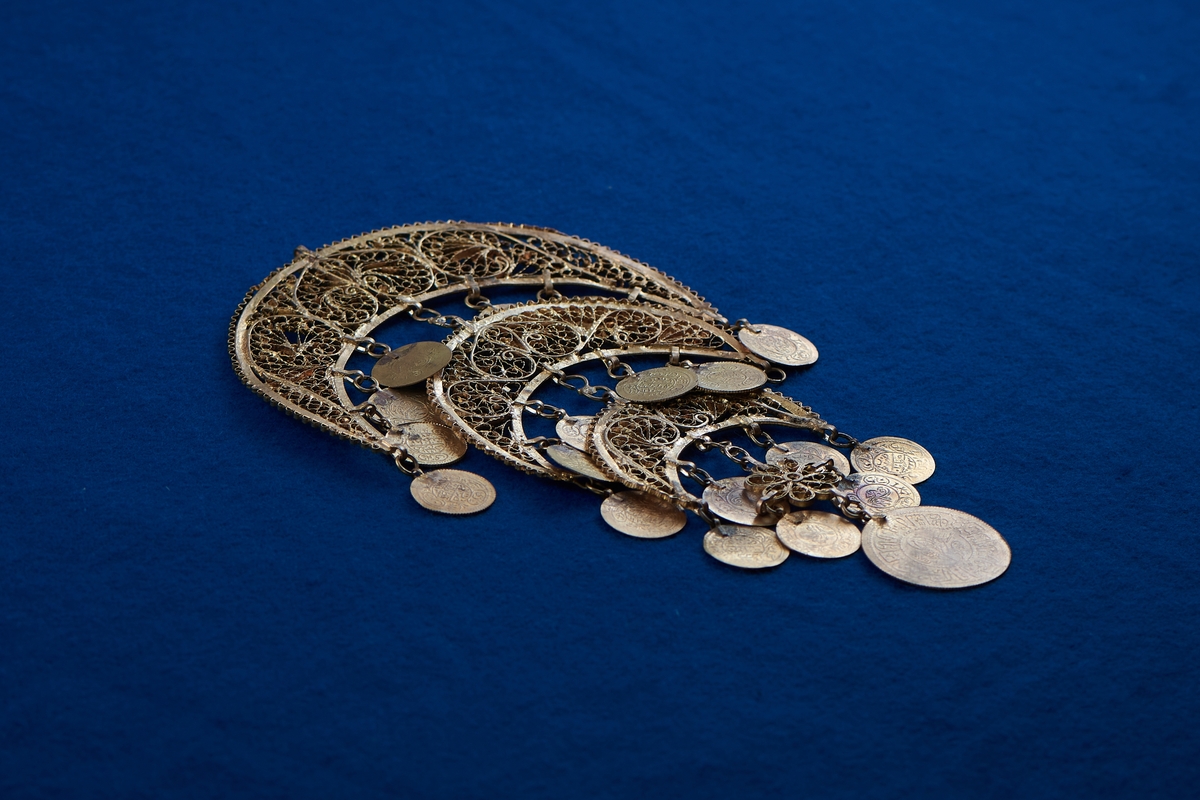 Del av halssmycke, bestående av metallbitar i form av tre halvmånar. Smycket är skapat i gul metall med infattade turkosfärgade stenar. Från varje halvmåne hänger cirklar i bindöglor. Smycket är troligen inköpt av Rosa Taikon vid en resa utomlands, och hängde senare på anslagstavlan i hennes ateljé.