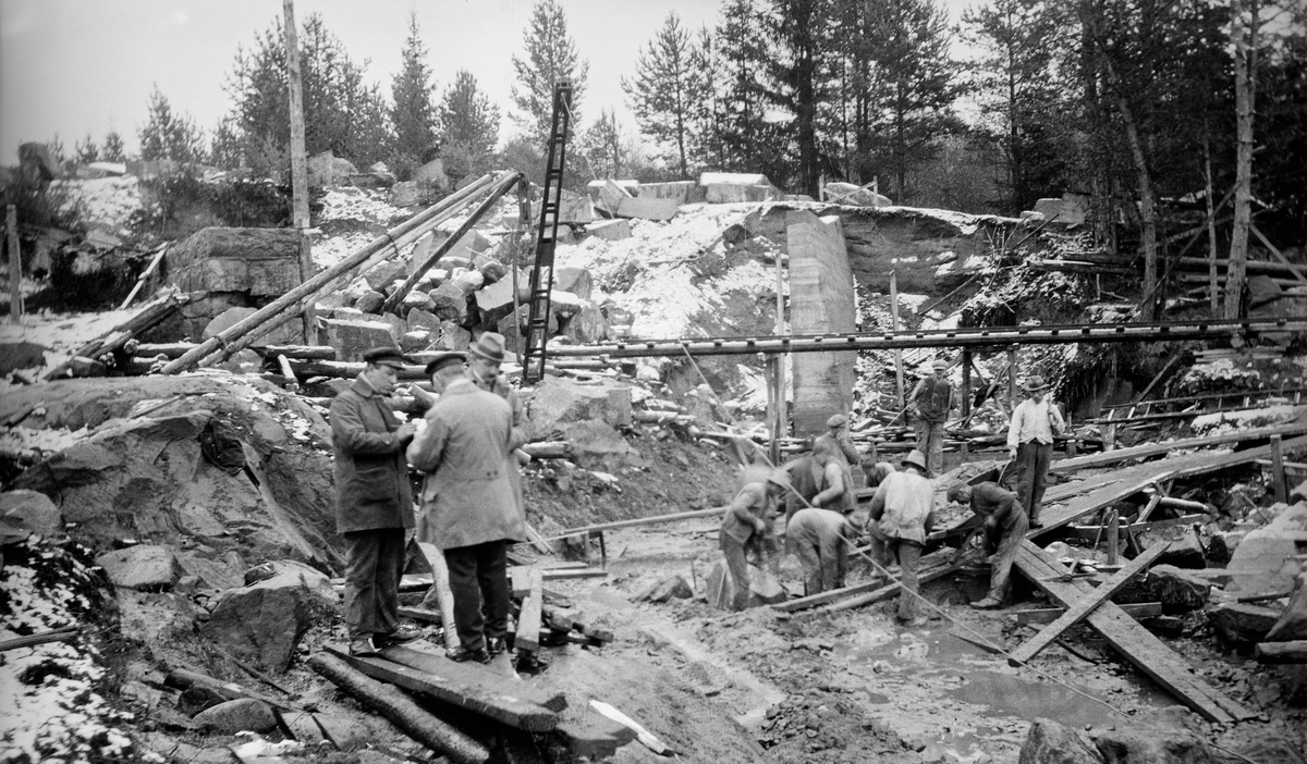 Fra ombygninga av Kjellåsdammen (Kjelåsfossdammen) i Flisa i Åsnes, Hedmark, i 1923. Fotografiet er tatt i anleggsområdet, der ni, muligens ti arbeidere hadde samlet seg rundt i grop i bakken. På et skråplan ved sida av dem sto det ei trillebår med grus eller muligens betong, som antakelig skulle tippes i gropa. Til venstre sto tre andre karer, den bakerste med hatt, frakk  og noe som kan ha vært en rull med tegninger i hendene, Bak dem ser vi ei kran og en kvadersteinsmurt mur. Lengre til høyre ser vi en støpt mur. På elvebrinken i bakgrunnen lå det en del digre, kantete steinblokker, som antakelig skulle brukes i den nye dammuren. Fotografiet er antakelig tatt en dag seinhøstes eller tidlig på våren da det lå et lite snødryss over terrenget.

Tidlig på 1900-tallet framsto Kjellåsdammen som et stort og kostbart fløtingsanlegg, som ble betjent av to damvoktere gjennom hele fløtingssesongen.  Hovedpoenget med anlegget var å magasinere vann, som kunne porsjoneres ut, slik at det ble mulig å få tømmeret videre nedover i retning Glomma også når vannstanden sank.  Effekten av dammen var omdiskutert, og enkelte mente at anlegget kunne fjernes, så snart det var utført litt mer mineringsarbeid i det nedenforliggende elveløpet.  I stedet bestemte fløtingsledelsen å bygge en ny og solidere Kjellåsdam, en murt steindam.  Arbeidet med denne konstruksjonen pågikk i perioden 1916-1924.  Kjellåsdammen var en såkalt attholdsdam, som skulle kunne brukes både til å heve vannspeilet i den ovenforliggende delen av elveløpet og til å regulere vannføringa i den nedenforliggende delen av Flisa-elva.