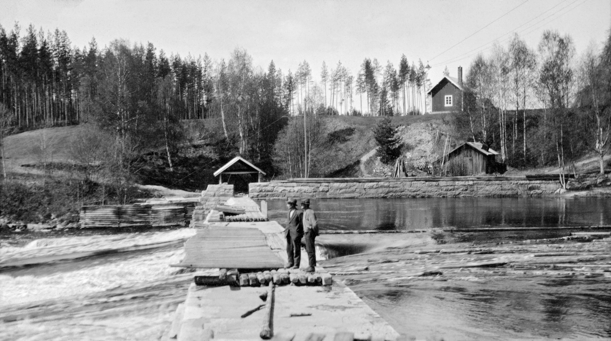 Fra Kjellåsdammen (Kjelåsfossdammen) i Flisavassdraget i Åsnes i Solør i Hedmark. Kjellåsdammen var en terskeldam, altså et stengsel på tvers av elveløpet som skulle stuve opp vann i det ovenforliggende vassdraget slik at elveløpet der ble djupere. På den måten reduserte konstruksjonen faren for at fløtingstømmer fra ovenforliggende skoger hektet seg fast på grus- og steinører i elveløpet. Dammen var bygd av kvadret naturstein som antakelig var forsterket med betong. Dammen hadde to løp. Det østre, nærmest fotografen, var delvis stengt ved hjelp av «nåler», vertikalstilte planker som var stilt tett i tett mellom en terskel på elvebotnen og dambrua på motstrøms side. Det vestre damløpet sto åpent. To menn befant seg på damkrona da dette fotografiet ble tatt, På motstrøms side, langs den vestre elvebredden ser vi en solid natursteinsmurt skådam som skulle hundre at tømmeret drev på land. På nedsida av dammen var denne forlenget med ei steinfylt tømmerkiste, Inne ved elvebredden sto det et skur og ei smie. Koia som skulle være innkvarteringssted for fløterne sto oppe på elvebrinken.