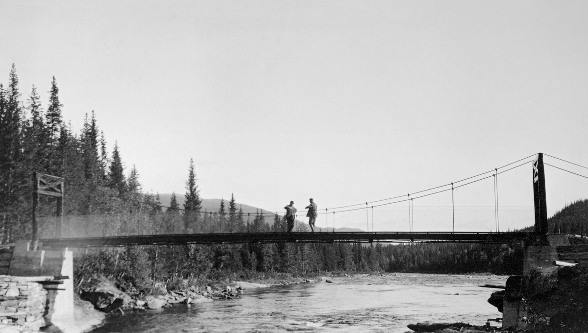 Den nye hengebrua - Fossbrua - over Atna i Stor-Elvdal, fotografert i 1930.  Fotografiet er tatt nedover vassdraget, og det viser hele bruspennet.  Brua er bæres av to trebukker som er plassert på avsatser foran på betongbrukar ved elveleiets ytterkanter.  I øverkant av brukaret til venstre i bildet er det murt en skjerm av naturstein, en skådam som skulle beskytte brukaret under flom og tømmerfløting.  Brulegemet bæres av jernstenger som er montert til vaierne og fastskrudd til tverrstilte jernbjelker på undersida av konstruksjonen. To menn står på brua.  Under brua går elva brei og stor.  Langs breddene vokser det granskog. 
