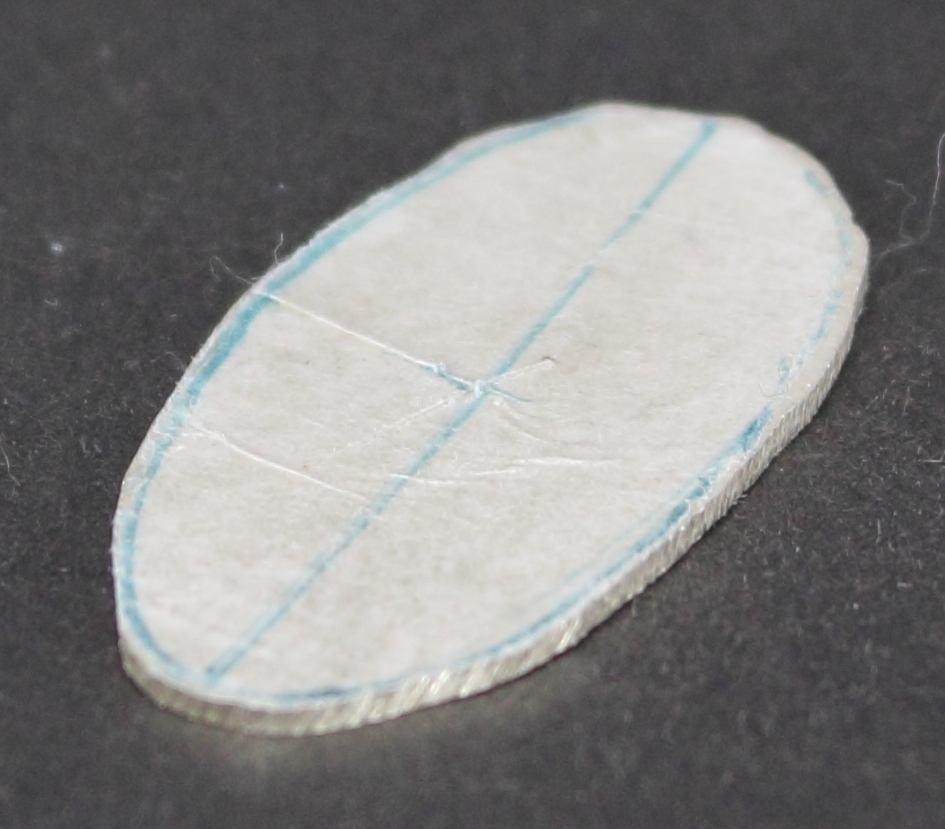 Silverplåt i oval form. På ena sidan finns papper fastlimmat. På pappret är streck ritade.  På andra sidan finns rester från avslitet papper.