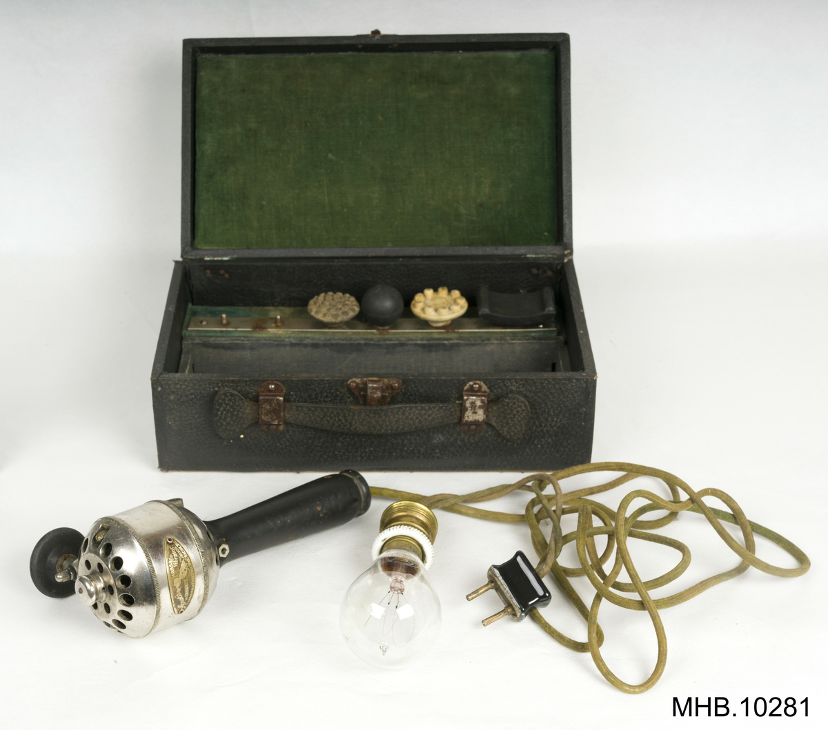 Massasjeapparat. Arnold Massage Vibrator,  Volt 110, med tilbehører i eske. 
Estetisk massasjeapparat med svart lakkert trehåndtak. Apparatet har en grønn kabel med svart porselen støpsel og glasslyspære (kulltrådslampe med spenning: 110V.)
Metallhodet inneholder motor, og rotor med et feste for tilbehører med utskiftbare elementer som to hoder i kremfarget gummi-latex og tre i svart bakelitt. 
Settet ligger inn i en rektangulær eske som en liten koffert grønn fløyelsfôret.