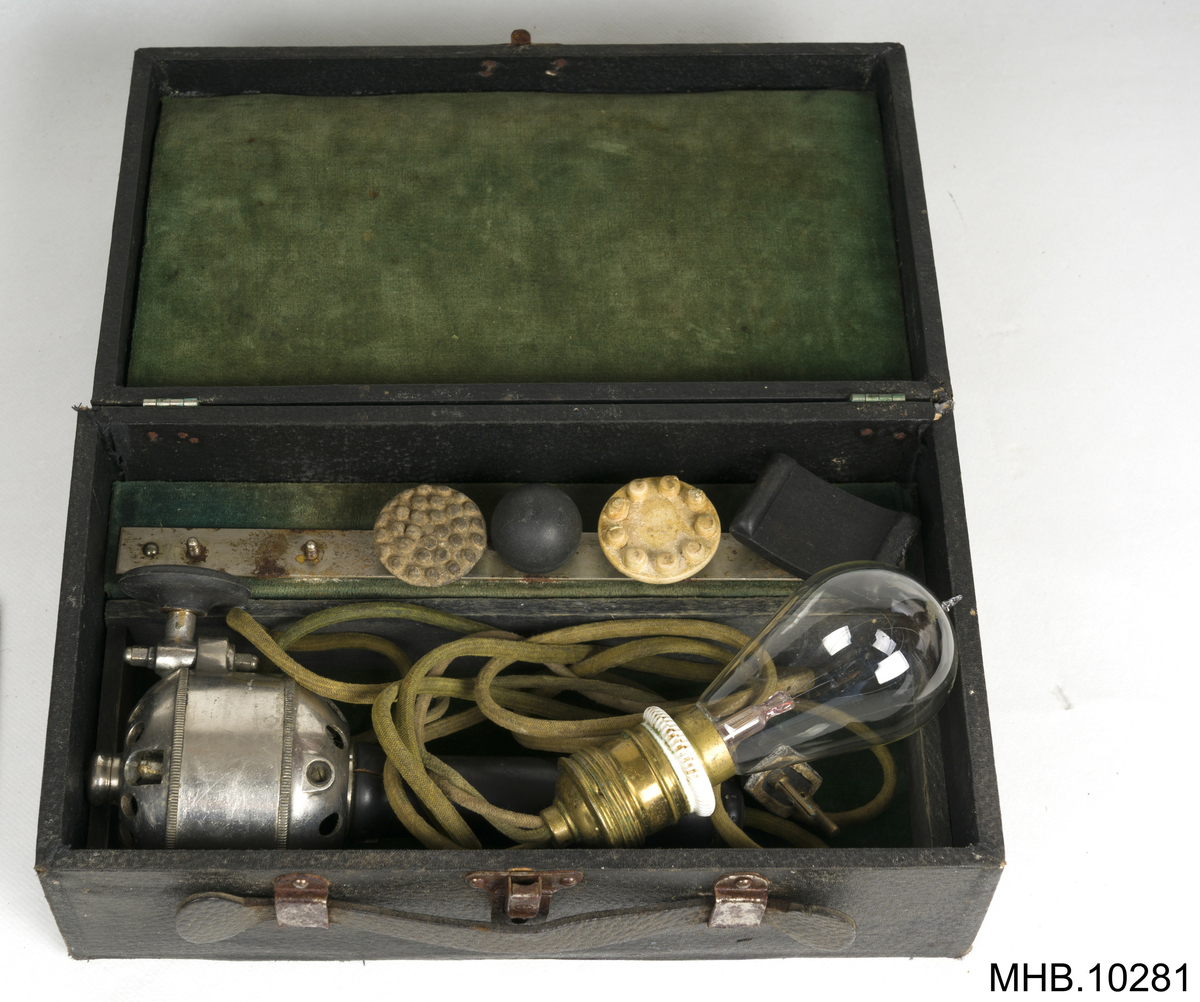 Massasjeapparat. Arnold Massage Vibrator,  Volt 110, med tilbehører i eske. 
Estetisk massasjeapparat med svart lakkert trehåndtak. Apparatet har en grønn kabel med svart porselen støpsel og glasslyspære (kulltrådslampe med spenning: 110V.)
Metallhodet inneholder motor, og rotor med et feste for tilbehører med utskiftbare elementer som to hoder i kremfarget gummi-latex og tre i svart bakelitt. 
Settet ligger inn i en rektangulær eske som en liten koffert grønn fløyelsfôret.