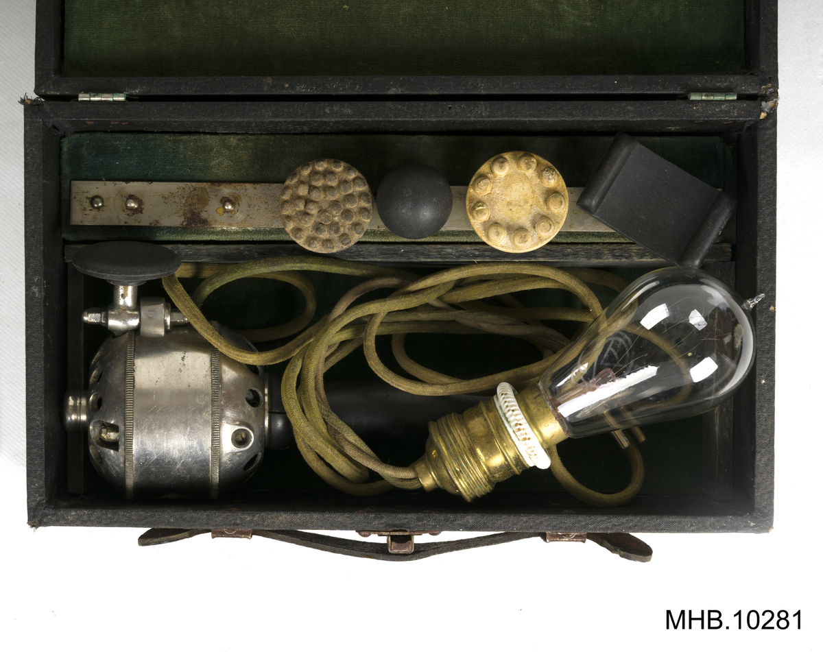Massasjeapparat. Arnold Massage Vibrator,  Volt 110, med tilbehører i eske. 
Estetisk massasjeapparat med svart lakkert trehåndtak. Apparatet har en grønn kabel med svart porselen støpsel og glasslyspære (kulltrådslampe med spenning: 110V.)
Metallhodet inneholder motor, og rotor med et feste for tilbehører med utskiftbare elementer som to hoder i kremfarget gummi-latex og tre i svart bakelitt. 
Settet ligger inn i en rektangulær eske som en liten koffert grønn fløyelsfôret.