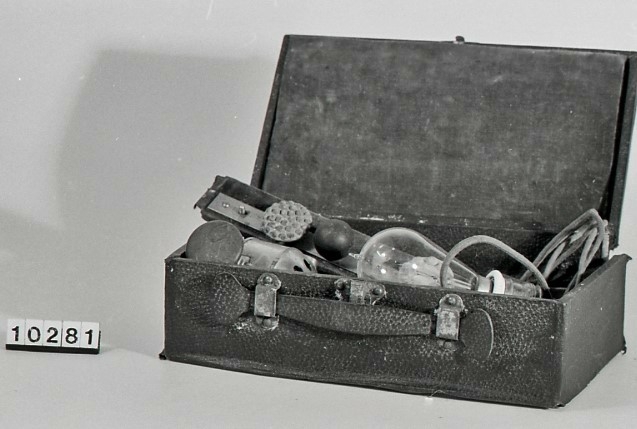 Massasjeapparat. Arnold Massage Vibrator,  Volt 110, med tilbehører i eske. 
Estetisk massasjeapparat med svart lakkert trehåndtak. Apparatet har en grønn kabel med svart porselen støpsel og glasslyspære (kulltrådslampe med spenning: 110V.)
Metallhodet inneholder motor, og rotor med et feste for tilbehører med utskiftbare elementer som to hoder i kremfarget gummi-latex og tre i svart bakelitt. 
Settet ligger inn i en rektangulær eske som en liten koffert grønn fløyelsfôret.