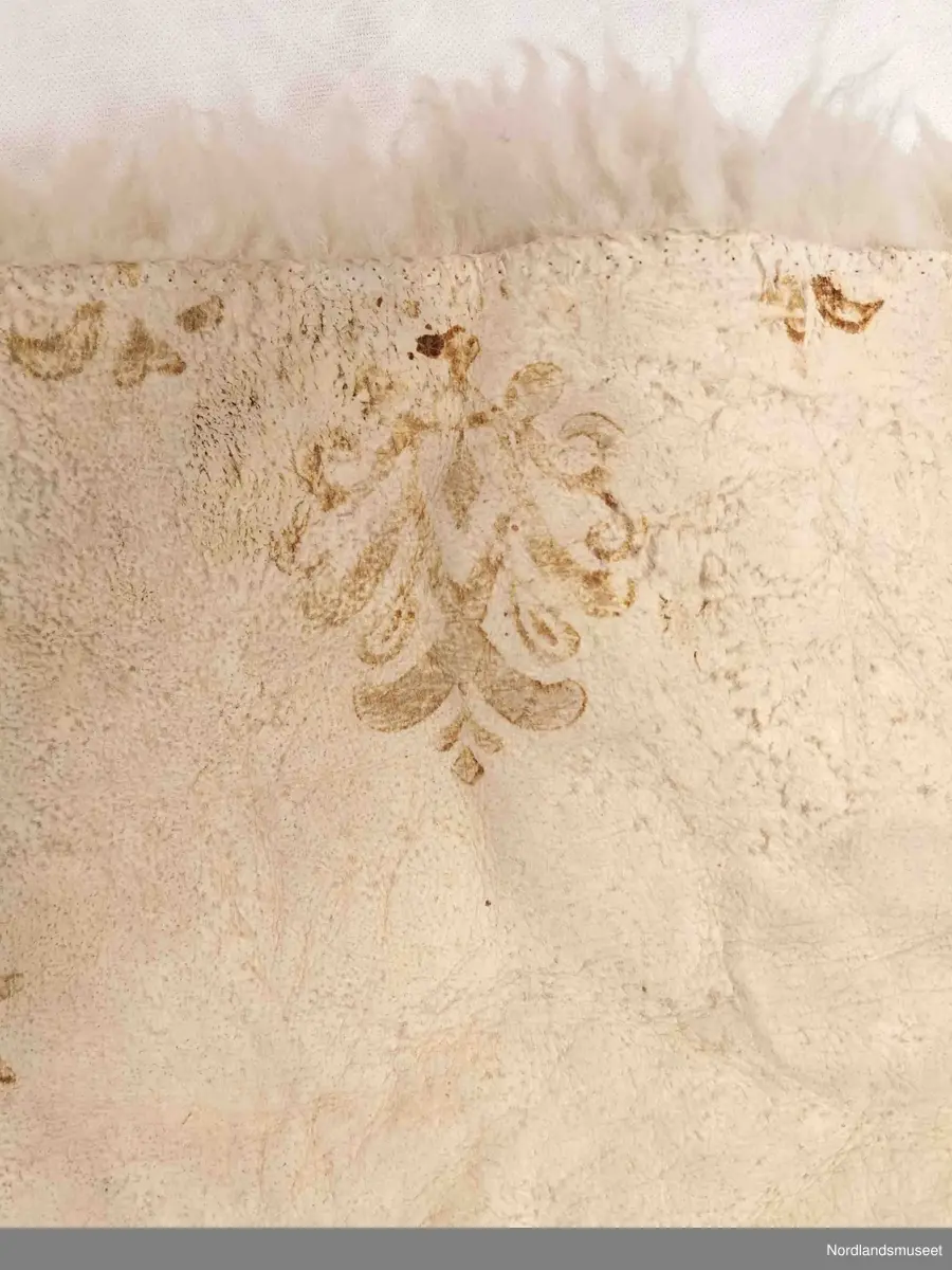 Rektangulær skinnfell laget av tre saueskinn sydd sammen. Fellen kan opprinnelig ha vært dobbelt så bred da den kun har kantbord i ene enden. Det er også rester av søm på den enden som mangler kantbord. 

Skinnfellen er dekorert med trykk av krinemønster. Her ser vi blant annet en hest.