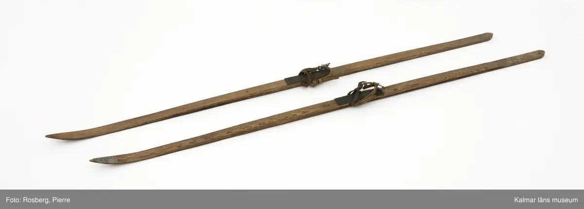 KLM 45695. Skidor, ett par. Av trä, mycket smala. Med gummiklädd fotdel. Bindning med läderremmar. Ena skidan lagad längst fram med fastspikad metallskoning.