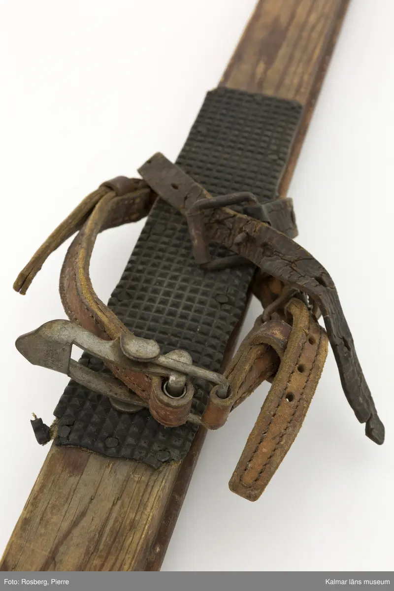 KLM 45695. Skidor, ett par. Av trä, mycket smala. Med gummiklädd fotdel. Bindning med läderremmar. Ena skidan lagad längst fram med fastspikad metallskoning.