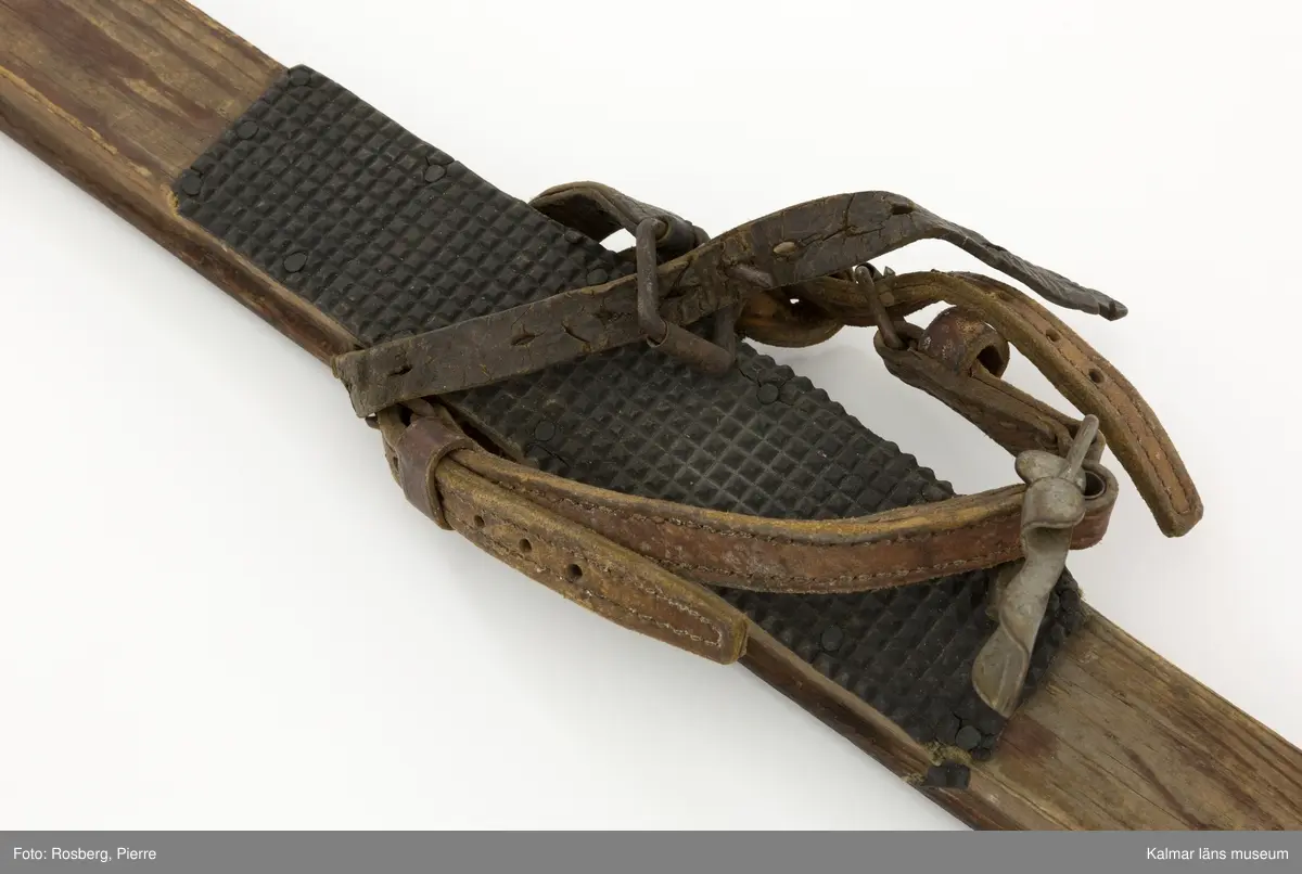 KLM 45695. Skidor, ett par. Av trä, mycket smala. Med gummiklädd fotdel. Bindning med läderremmar. Ena skidan lagad längst fram med fastspikad metallskoning.