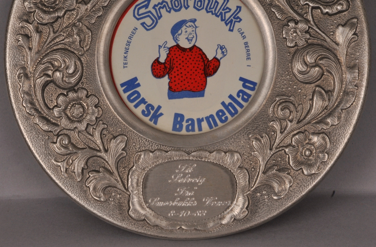 Plakett frå Smørbukks vener. Rund metallramme med roserankar kring kanten. Teksten:"Til Solveig frå Smørbukks vener 8-10-83". Bilete av Smørbukk med teksten "Teikneserien Smørbukk går berre i Norsk Barneblad"  i midten av ramma.