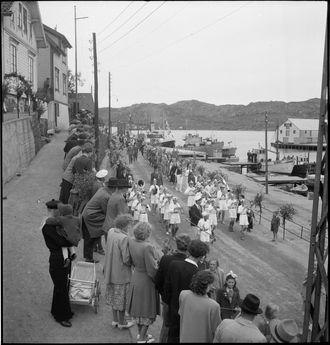 Folketog på 17. mai. Flere korps og barn går langs Vågen og igjennom sentrum av Egersund. Mange har kommet for å se toget.