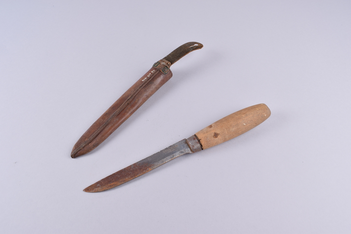 Kniv med skaft av ubehandlet tre og slire i lær.
Stålblad og -holk. Sliren har en bærestropp av sort lær med knapphull.