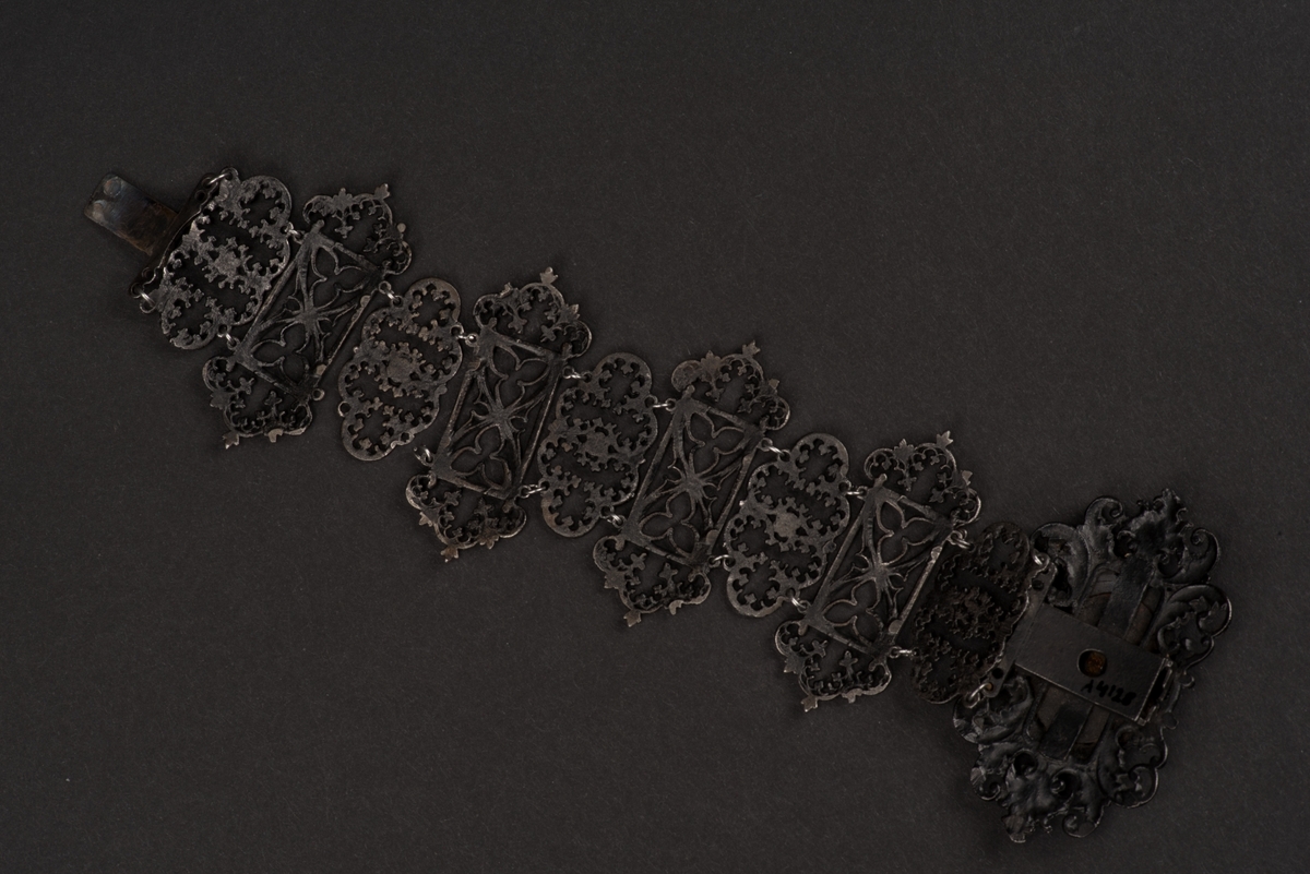 Svart armband av tio ledade länkar av gjutjärn.
Länkarna är utformade som gotiska trepass dekorerade med växtornament i rokokostil. Länken som döljer låset är större än de övriga och dekorerad med ett sittande figur som framhävs genom en förnicklad plåt under.