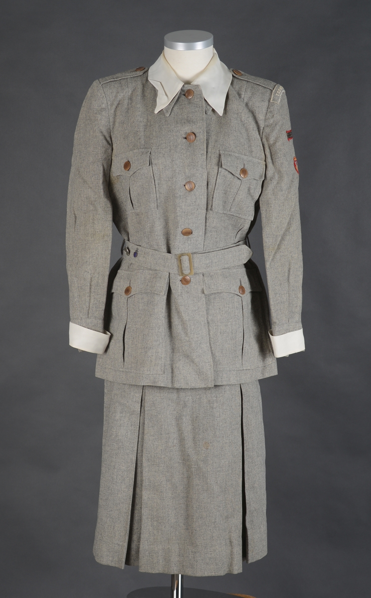 Lotteuniform består av jakke, skjørt og belte. Enkeltknept uniforms jakke med 4 utenpåsydde lommer med motfold og klaffer som lukkes med knapp og knapphull. Jakken lukkes med 1 trykk-knapp, 5 knapper og knapphull. 2 innsnitt i livet og 1 innsnitt fra skulder ned til bysten på hvert framstykke. Bakstykket har 4 innsnitt fra overkant av livlinjen og ned til nedre kant av jakken. Bærestykke øverst på ryggen. Ryggstykket er svakt rynket til bærestykket. Skulderstropper som festes med knapp og knapphull. Lange ermer med liten splitt og mansjett. Ermene er lagt i legg mot mansjettene. Jakken har skulderputer og innlegg under ermhulen.  Øverst på venstre erme er det sydd på 3 merker. Det øverste merket er av draktstoffet med tekst: Trondheim. Under det er det et rektangulært merke. Nederst et rundt merke, NLF merke. Løse hvite mansjetter på erme og løs krage rundt halsen. Kragen er sydd fast for hånd med lange sting. Jakken er lagt opp for hånd. Jakken har belte med spenne i livet og 2 beltestropper som er festet i sidene. På beltet er det to merker, 1 rundt sølvfarget festet med nål og et merke som er sydd fast.
Skjørtet har 2 motfolder foran og 2 bak. Linning i livet og åpning i venstre side som lukkes med metallglidelås, knapp og knapphull. Skjørtet er lagt opp for hånd. Drakten er uforet. Merkelapp langs glidelåset med tekst. 
Alle knapper på uniformen er merket NLF.