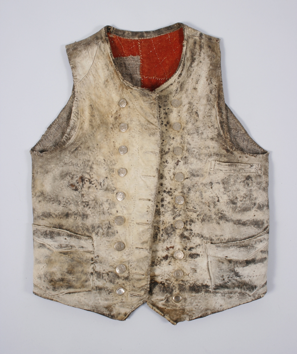 Dobbeltknept skinnvest med 9 knapper og passepoilerte knapphull på hvert framstykke. Knappene er festet med en skinnreim på innsiden . Den er sydd fast til foret. Øverste knapphull på høyre framstykke er sydd med knapphullsting. Knappene er siselerte med bokstaven B i midten med en bord rundt.  Den nederste knappen på begge framstykkene er mindre enn de andre og har ikke samme mønster som de andre. Vesten har 1 brystlomme og 2 lommer i livhøyde. 1 innerlomme i skinn er sydd fast til foret. Foret er sydd sammen av mange små stoffbiter i 2 farger. Foret og skinnet er stukket sammen. Bakovertrekte sidesømmer. Påsydd skinnkant rundt halsrigning og langs begge sider MF. Framstykkene går ned i en spiss MF.