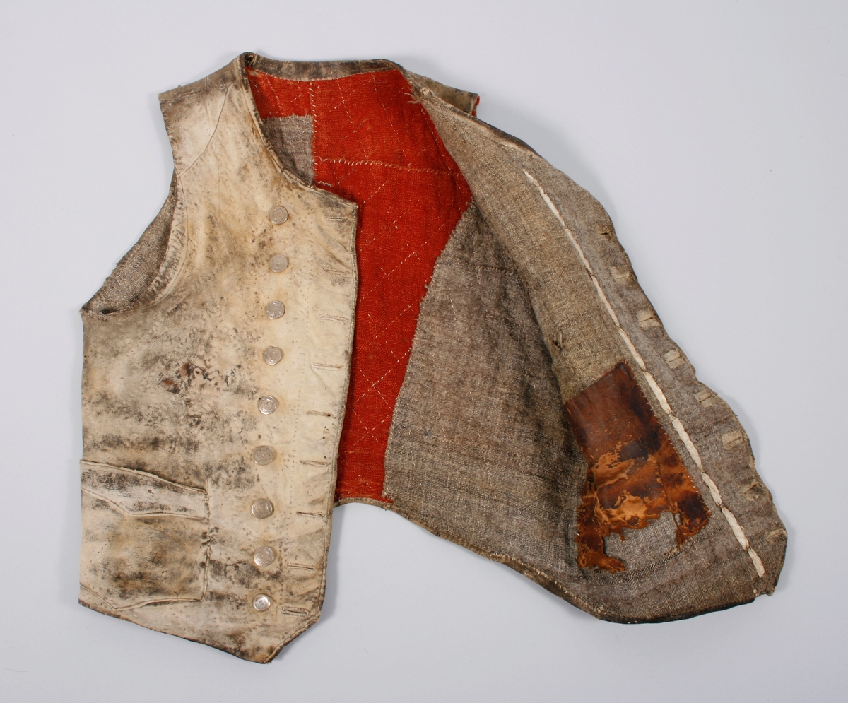 Dobbeltknept skinnvest med 9 knapper og passepoilerte knapphull på hvert framstykke. Knappene er festet med en skinnreim på innsiden . Den er sydd fast til foret. Øverste knapphull på høyre framstykke er sydd med knapphullsting. Knappene er siselerte med bokstaven B i midten med en bord rundt.  Den nederste knappen på begge framstykkene er mindre enn de andre og har ikke samme mønster som de andre. Vesten har 1 brystlomme og 2 lommer i livhøyde. 1 innerlomme i skinn er sydd fast til foret. Foret er sydd sammen av mange små stoffbiter i 2 farger. Foret og skinnet er stukket sammen. Bakovertrekte sidesømmer. Påsydd skinnkant rundt halsrigning og langs begge sider MF. Framstykkene går ned i en spiss MF.