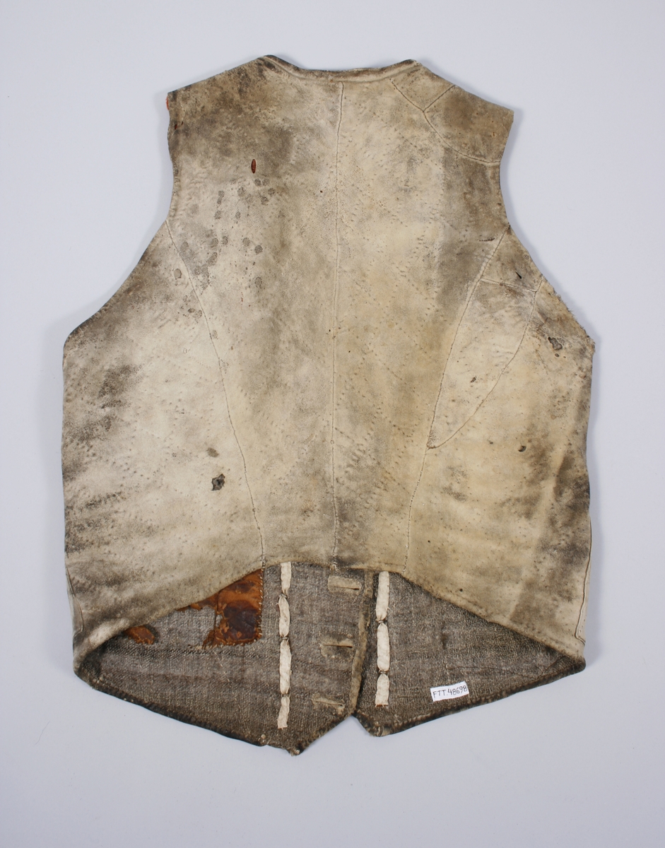 Dobbeltknept skinnvest med 9 knapper og passepoilerte knapphull på hvert framstykke. Knappene er festet med en skinnreim på innsiden . Den er sydd fast til foret. Øverste knapphull på høyre framstykke er sydd med knapphullsting. Knappene er siselerte med bokstaven B i midten med en bord rundt.  Den nederste knappen på begge framstykkene er mindre enn de andre og har ikke samme mønster som de andre. Vesten har 1 brystlomme og 2 lommer i livhøyde. 1 innerlomme i skinn er sydd fast til foret. Foret er sydd sammen av mange små stoffbiter i 2 farger. Foret og skinnet er stukket sammen. Bakovertrekte sidesømmer. Påsydd skinnkant rundt halsrigning og langs begge sider MF. Framstykkene går ned i en spiss MF.