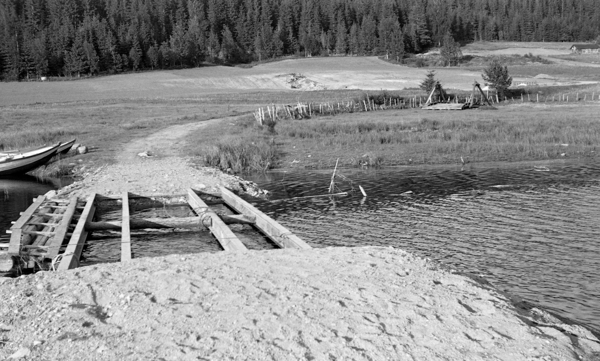 Bjelker for ny bru over bekken Størja ved Jersås i Sør-Odal i Hedmark. Fotografiet er tatt fra vegtraseen mot krysningspunktet ved bekken, der dert var lagt H-bjelker av stål, med mellomliggende kabber av rundtømmer som antakelig skulle låse den innbyrdes avstanden mellom bjelkene. Oppå skulle det antakelig legges en bane - muligens av kraftige planker - som kunne bære de kjøretøyene som skulle passere brua. Høyden fra vannspeilet opp til bjelkene var bemerkelsesverdig liten. På motsatt side av bekken var det et åker- og englandskap, som etter hvert ble avløst av ei skogli. Til venstre i forgrunnen, ved bekken, skimter vi stevnene på to robåter.