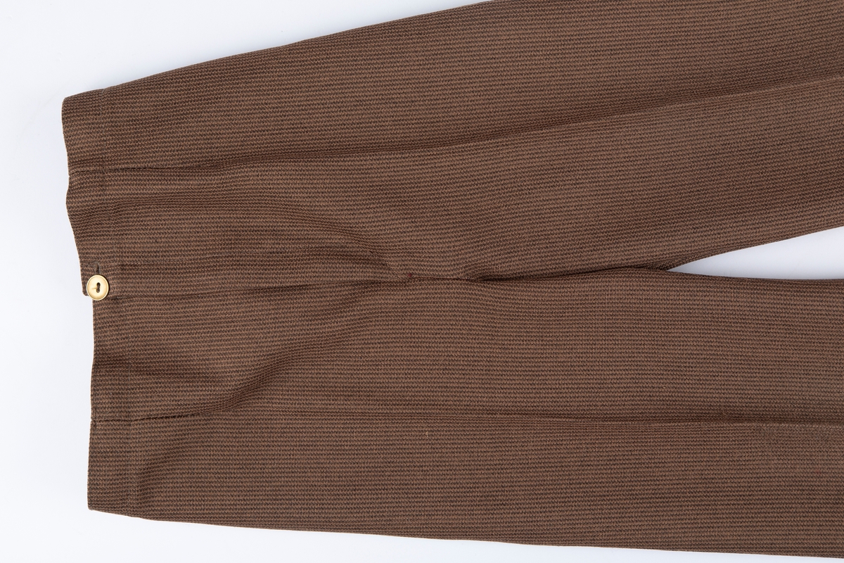 Buksedress. A: Jakke. B: Bukse.

A: Jakke. 
Rund hals, uten krage. 3 par messingknapper, 3 knapphull. Buet lengdesnitt foran og bak. Lange ermer. Falske lommeklaffer i snittsøm foran.

B: Bukse: 
Nedbrettet linning, med strikk på bakstykket. Glidelås i gylf. Buksebena har "sleng". 
Buksen har produksjonsmerke for stoffet