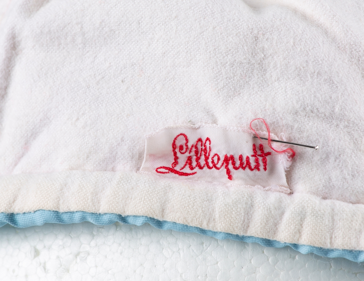 Hjelm med dusk og brem. Foret med hvitt flanellstoff. Strikket kant i front. Løpegang i nakke med knyttebånd. Fabr. Merket "Lilleputt".