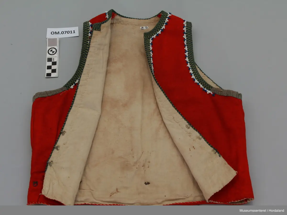 Raudt ull-liv med gul og grøn krokakvarde i silke. Anna bandbit under armane. Breid perlerad innanfor med kvite og blåe perler. Livet har innsnitt nede, midt på kvart framstykke. Hekter utanpå livet nede, rundt heile livet, for å hekta på stakk. Tre hekter framme til attlat. Bomullsfor. Handsaum.