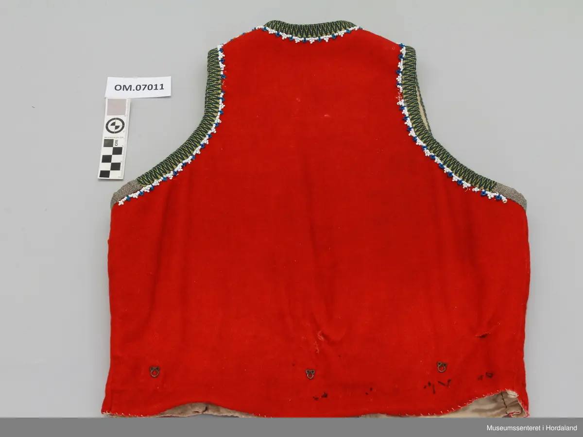 Raudt ull-liv med gul og grøn krokakvarde i silke. Anna bandbit under armane. Breid perlerad innanfor med kvite og blåe perler. Livet har innsnitt nede, midt på kvart framstykke. Hekter utanpå livet nede, rundt heile livet, for å hekta på stakk. Tre hekter framme til attlat. Bomullsfor. Handsaum.