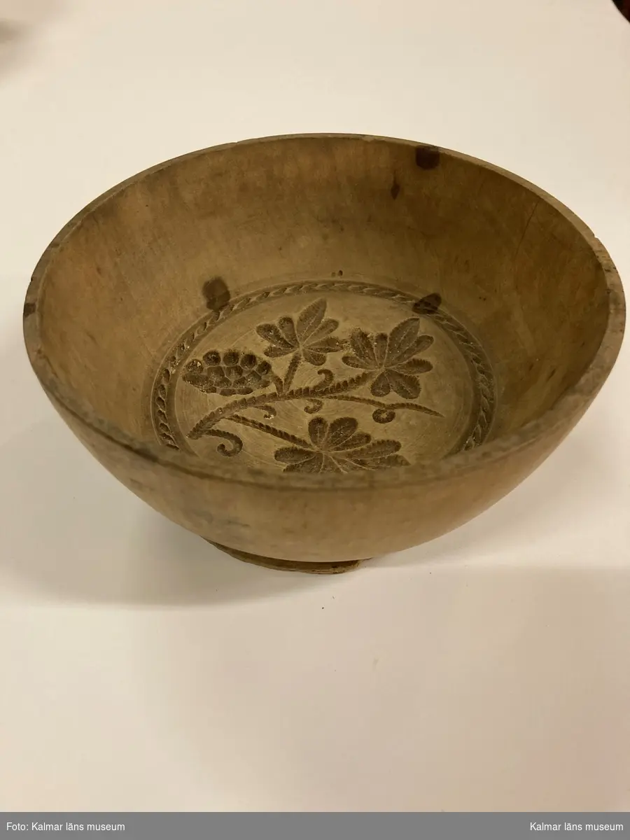 KLM 6153. Ostform. Av trä. Svarvad skål med låg fot. I botten ett snidat mönster i form av en vinkvist med en druvklase.